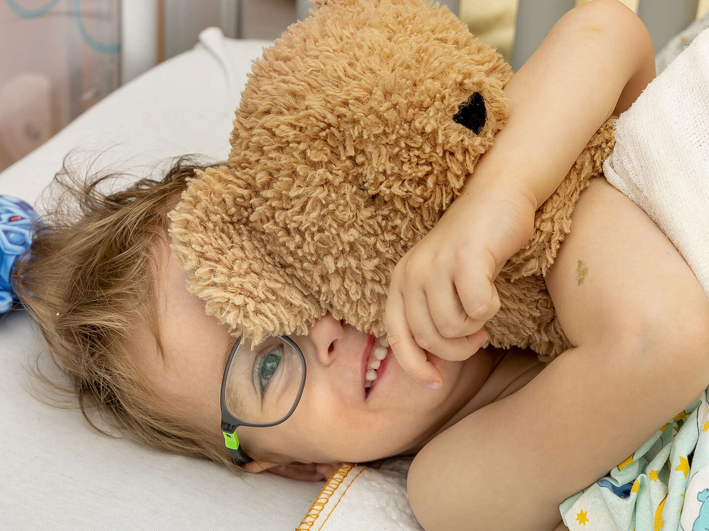 Kind im Bett liegend mit einem braunen Teddybär im Arm.